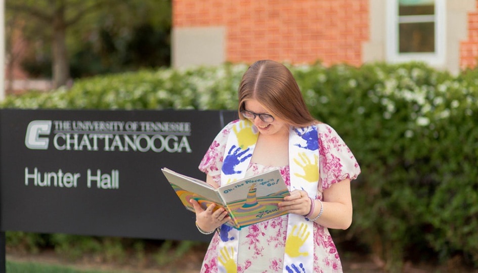 Graduate, Kendell Wedekind, holding a book in front of Hunter Hall.