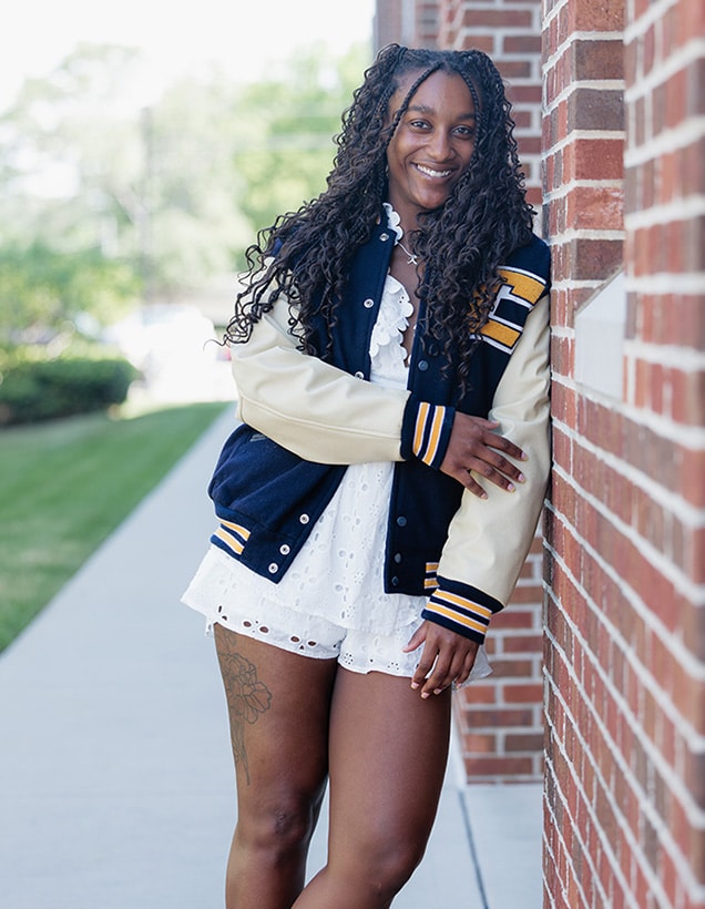 Kalifa Ford wearing a navy-and-gold letterman jacket over a white eyelet romper, standing beside a red brick wall on a sidewalk.